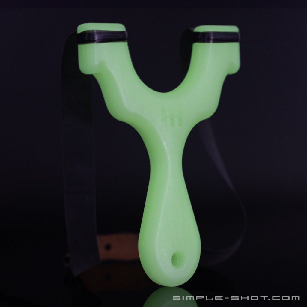 A Glow-in-the-Dark Axiom Champ Slingshot with a sleek, modern design, featuring a Y-shaped frame and black elastic bands. The handle boasts a textured grip for superior hold, and the minimalist gear brand name 