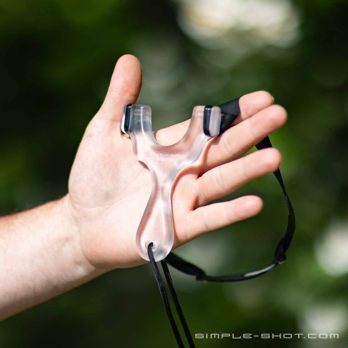 A hand is holding a compact Axiom Champ slingshot with a black lanyard attached. The background consists of blurred greenery, creating a natural outdoor setting. The product appears sleek and ergonomically designed. The words "SIMPLE-SHOT.COM" are visible in the bottom right corner.